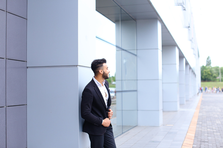 Young handsome businessman Arab Muslims is in the business center in black elegant dress on building background. Businessman man male looking at time walking goes and smiling.の写真素材