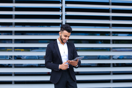 Entrepreneur uses tablet for online order shopping with debit credit card looking smiling at the camera. Young handsome businessman arab muslims in business center wearing dressed in black elegant suit on building background.の写真素材