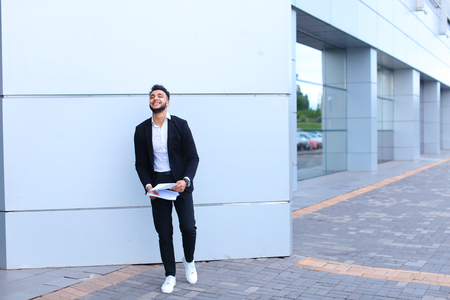 Young beautiful male guy arab businessman student laughing, smiling, rejoices success, holds papers or documents, stands in full growth directly into camera against background of gray wall of large shopping business center. Muscular bearded man dressed inの写真素材