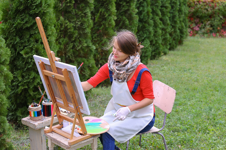 Female artist holds brush and wrote oil on canvas painting, does what likes, interests and hobbies,  preparing picture to order, advertises or art studio for drawing utensils, sits on chair at  wooden easel in large green park outdoors. Girl with brown haの写真素材