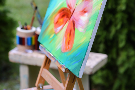 Photo completed and unfinished picture, works of art on which  image multi-colored butterfly on canvas and stands on wooden easel, palette and brush lying near easel, on table stands colorful banks in which there are other artistic intrumenty and colored の写真素材
