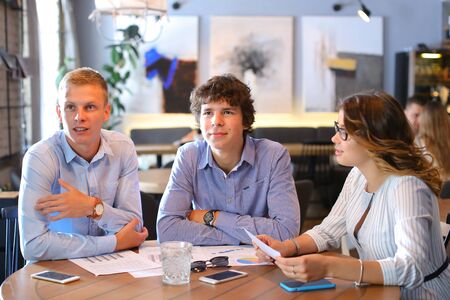 Two smart young business men one woman in suits sitting on table with documents, graphics, technology, laptop, phone, tablet. Team entrepreneurs negotiate, discuss issues, solve problems, agree, talkの写真素材