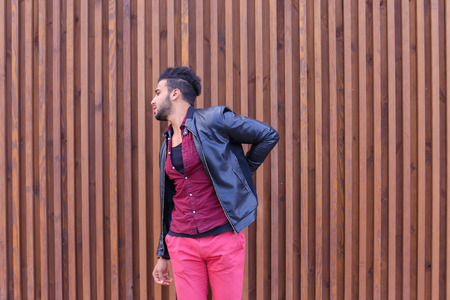 Handsome Young Muslim Man, Businessman Suffering From Back Pain, Stretching, Trying to Warm up and Stands on Background of Wooden Panel Stairs Outdoors. Attractive Arabic Bearded Man With Dark Hair Dressed in Black T-Shirt Over Maroon Shirt and Wears Blacの写真素材