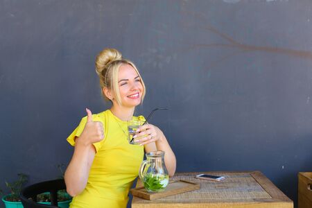 Stunning cute young girl female woman looks forward, smiling broadly and shows gesture thumbs up,  holds glass in hand, looks at camera, laughing and sits at wooden square table against background of gray wall in coffee shop outdoors. Girl with blond hairの写真素材