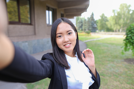 Portrait of young asian businesswoman female uses cellphone takes selfie on frontal camera on background on offices and trees in open air. Concept of International Multicultural Businesses Work Relations, Technology, Modern Entrepreneur, selfie.の写真素材