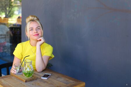 Stunning cute young girl female woman smiling, holds glass in hand, looks at camera and sits at wooden square table against background of gray walls and windows in coffee shop outdoors. Girl with blond hair dressed in short citric dress.Concept of meetingの写真素材