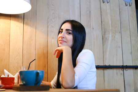 Pretty cute young female woman smiling, posing and looking in camera at wooden table on background of wooden wall panel in cozy coffee shop. Girl with black hair dressed in white sweater. On table stands desserts, coffee cups and glass cup with lemonade. の写真素材