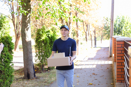 Messenger Carring Two Boxes in Hands Smiling on Background of Trees Along Street. Courier Company Provides Its Services in All Areas Ofterrain, Man of European Appearance in Uniform Carries Two Boxes With Order Along Street on Background of Shrubs, Trees の写真素材