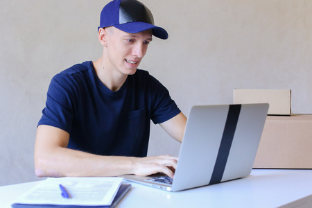 Young Postman Sits at Table Typing at Laptop. Courier Company Accepts Orders Every Day Different Types and Sizes Young Deliveryman European Appearance in Black T-Shirt and Cap Sitting at White Table Typing on Computer and Takes Pen Writing Down Informatioの写真素材