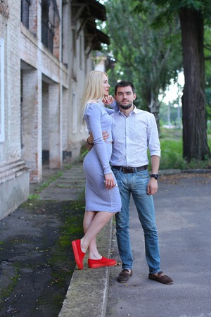 Young couple standing next to each other, posing and hugging on street. Girl turned sideways in profile and dressed in midi gray dress and red slip-on, guy standing straight and looks straight into camera, he is dressed in blue jeans and white striped shiの写真素材