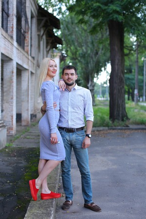 Young couple standing next to each other, posing and hugging on street. Girl turned sideways in profile and dressed in midi gray dress and red slip-on, guy standing straight and looks straight into camera, he is dressed in blue jeans and white striped shiの写真素材