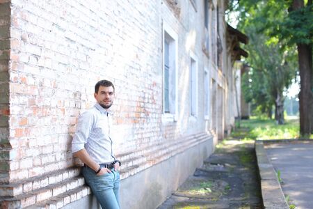 Young handsome guy looking into camera, standing near stone brick gray wall in street and holding hands in pockets of pants. Man dressed in white shirt with red stripe and blue jeans. Concept of business man waiting for meeting, posing in front of camera,の写真素材