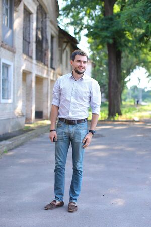 Handsome male man guy smiling and standing still on street concrete road. Young man dressed in white shirt with red stripe, blue jeans and brown loafers. Concept of business man waiting for meeting, posing in front of camera, casual style.の写真素材