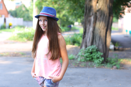 Beautiful girl child smiling, looking into camera, with long hair standing in yard on street on background of trees and houses. Girl dressed in pink shirt, printed shorts, dark blue hat with a brim. Concept advertising of childrens clothes, walk and summeの写真素材