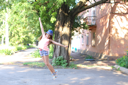 Pretty cute female girl child jumping, fun, fooling around and laughing in yard on street summer day. Girl dressed in pink shirt, printed shorts, dark blue hat with brim and blue sandals. Concept of fun, children happiness, childhood, summer holidays.の写真素材