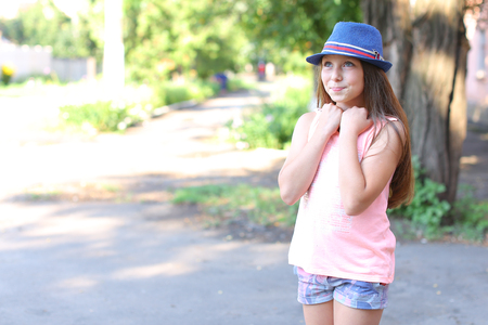 Pretty cute female girl child smiles, make wish, dreamily looking away, holding hands near face and standing in yard on street. Girl dressed in pink shirt, printed shorts, dark blue hat with brim. Concept of childhood, desire, dream, summer holidays, goodの写真素材