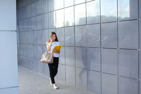 Serious young adult business woman walks and talks on phone, solves problems, holds and looks at documents in yellow folder on background of gray wall of shopping business center. Girl dressed in white shirt, black pants, beige shoes and keeps on hand beiの写真素材