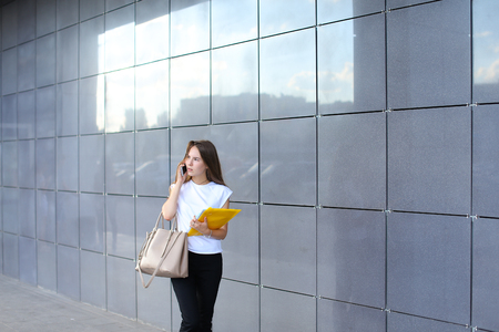 Serious young adult business female woman talks on phone, negotiating and looks aside, holds documents in yellow folder on background of gray wall of shopping business center. Girl dressed in white shirt, black pants and keeps on hand beige bag. Concept oの写真素材