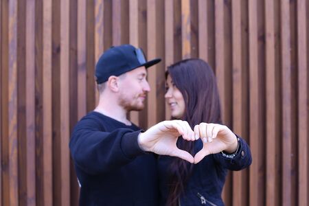 Romantic man and woman show heart in camera, kissing and cuddling, looking at each other and in camera, enjoying life together. Guy dressed in black sweater, blue jeans, sneakers and black cap, girl with long black hair dressed in black sweater, blue jeanの写真素材