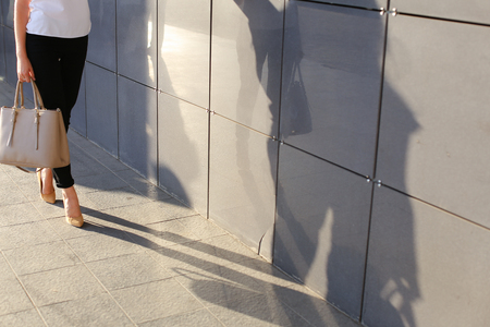 Photo legs of girl who goes forward on gray tile road against background of gray wall of modern business center. Girl dressed in white shirt, black classic trousers and beige high heels, big beige bag in hand. Concept of advertising, advertising clothes, の写真素材