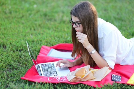female in the park works behind laptop. beautiful girl with glasses eating fast food in nature and smiling.の写真素材
