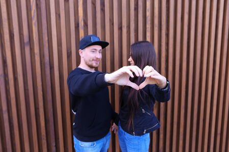 Romantic man and woman show heart in camera, kissing and cuddling, looking at each other and in camera, enjoying life together. Guy dressed in black sweater, blue jeans, sneakers and black cap, girl with long black hair dressed in black sweater, blue jeanの写真素材
