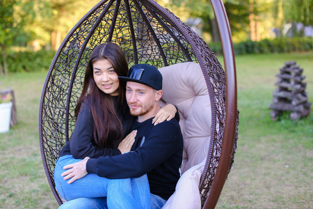 Couple in love swinging on chair in street among  large green park, discussing plans for future, share secrets and jokes, young people hugging each other gently. Guy dressed in black sweater, blue jeans, sneakers and black cap, girl with long black hair dの写真素材
