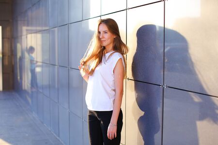 Young cute female girl student smiling, straightens hair, posing for camera and stands near modern business cente. Girl with long light brown hair dressed in white shirt and black classic pants. Concept of student everyday life, work week, homework, studyの写真素材