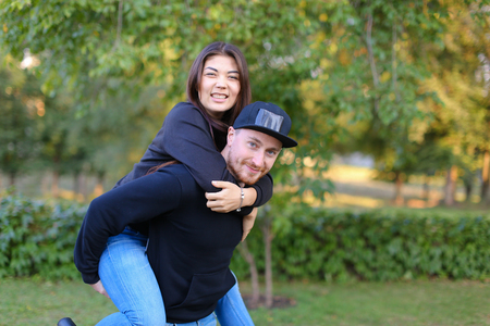 Pair of young fool around and have fun, fall into childhood and fun leisure time together, she rides guy in love joking and laughing, posing for camera in beautiful large green park outdoors. Man dressed in black sweater, blue jeans and black cap, girl wiの写真素材