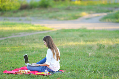 young girl sitting in the park and talking with headphones through the notebook. student speaks with parents rodnvmi with friends in nature.の写真素材