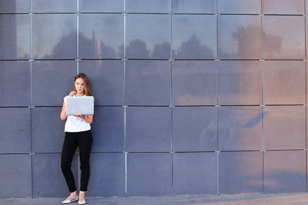 Promising young entrepreneur beautiful woman uses laptop for work, make appointment, solves important business issues and and stands right in all growth against the background of gray wall of modern business center. Girl dressed in white shirt, black pantの写真素材
