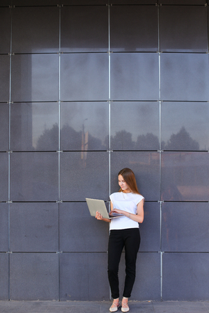 Promising young entrepreneur beautiful woman uses laptop for work, make appointment, solves important business issues and and stands right in all growth against the background of gray wall of modern business center. Girl dressed in white shirt, black pantの写真素材