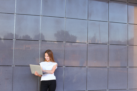 Beautiful young female woman girl entrepreneur successful person holds laptop in hands works, solves problems, and stands on background wall of modern business center.Girl with long hait dressed in white shirt and black trousers.Concept of business meetinの写真素材