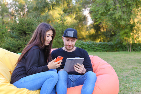 Pair of lovers of young attractive people sitting in soft yellow and red chair, man uses gadget that keeps in arms, female holds plastic card, coupon. Young people cute communicate, plan to travel, choosing interior of apartment, she shows on flyer and guの写真素材