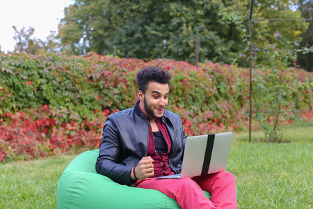 Perspective Young Arabic Guy, Businessman, Student Sits With Laptop in Soft Green Chair, Looks at Computer Screen and Smiling, Rejoice Success, Winning on Stock Exchange, Working Online on Background of Green Grass in Garden Outdoors. Bearded Man Dressed の写真素材