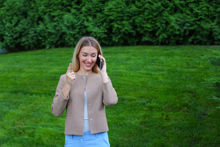 Young modern woman chatters with friend on smartphone, tells funny story and jokes, listens attentively interlocutor at other end of line, shares secrets, shows emotion of joy and gestures. Blonde with long hair dressed in light blouse, beige jacket and bの写真素材