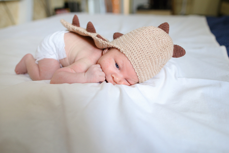 Newborn boy in funny brown knit dragon suit lies on white bed in bedroom. Concept of Maternal Love, Affection, Beginning Life, Cognition Surrounding World.の写真素材