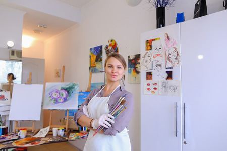 Cute female painter posing, laughing and fooling around in front of camera and smiling with brushes and colorful palette in hand, standing in studio with white art paintings on walls. Girl European appearance advertises art school for adults and children,の写真素材