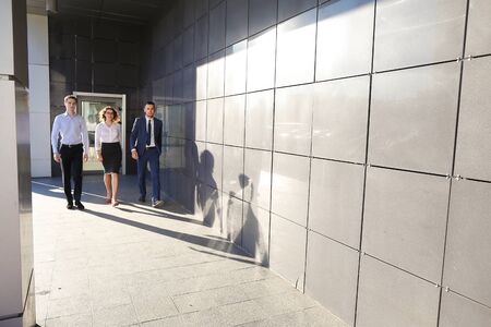Beautiful successful energetic people, two guys and girl, young businessmen, students confident step forward, smiling and looking at camera on background of gray wall outdoors business center. One of guys dressed in blue classic suit, white shirt with tieの写真素材
