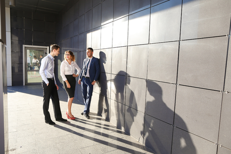Contemporary perspective elegant young people, two boys and a girl, businessmen and students to meet and discuss important issues, share business ideas, smiling, laughing, make decisions, solve problems and standing near gray wall of modern business centeの写真素材