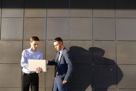 Handsome confident young male man, businessman, student holds laptop in hands and deliver presentations, talks about gadget, advertises new technology to colleague who listening to attentively, and stand on background wall of modern business center outdooの写真素材