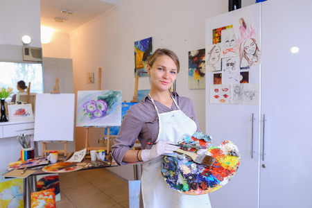 Cute female painter posing, laughing and fooling around in front of camera and smiling with brushes and colorful palette in hand, standing in studio with white art paintings on walls. Girl European appearance advertises art school for adults and children,の写真素材