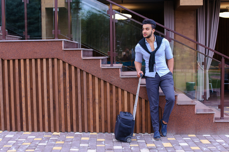 Young man businessman Arabian beautiful appearance is black travel bag on small wheels and is waiting for taxi, on journey or business trip, one leg held up other, and based on stairs near restaurant on open air. Handsome Muscular Guy Dressed in Black Claの写真素材
