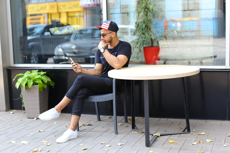 Young handsome arabic guy uses phone, sits chatting and smiles, holds hand near face, sitting on wicker gray chair at table on background of panoramic windows and flowers in modern restaurant, coffee shop. Man dressed in dark T-shirt, black pants, white sの写真素材