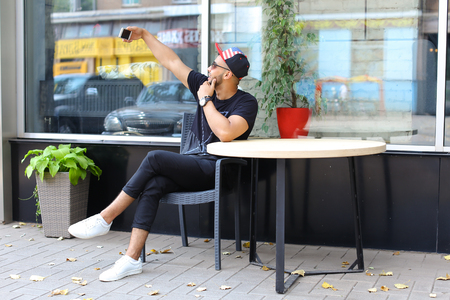 Lovely masculine Arabic guy makes selfie, raising arm and sits in gray wicker chair, smiles on background of panoramic windows in modern restaurant. Man dressed in dark T-shirt, black pants, black color red cap and sunglasses. Concept of selfie, in Skype の写真素材