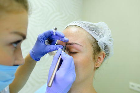 Successful female browmaker consult client girl completes registration and drawing eyebrow hairs and antiseptic wipes. Master dressed in white robe, blue rubber gloves and face protection gauze bandage. Concept of mikrobleyding eyebrows micropigmentation の写真素材