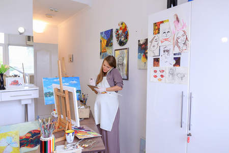 Lovely Female Painter Orders Things to Draw or Picture Frames , Clarifies Meeting With Students For Master Class, Communicates With Clients About Sale of Paintings. Woman With Long Light Brown Hair Dressed Sht Pale Purple Jacket, White Apron and White Gloの写真素材