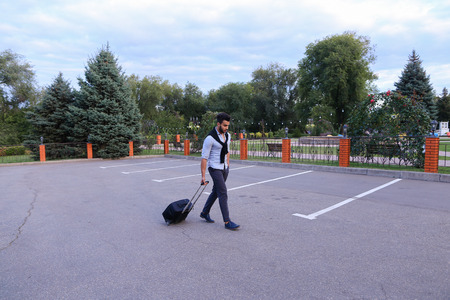 In Parking Goes Young Man Arabic Attractive Appearance With Beard and Dark Hair Dressed in Suit Black Pants White Shirt With Black Sweater on Shoulders and Roll Black Suitcase Laggage in His Hand. Concept of Business Trip, Business Meeting, Travel, Busineの写真素材