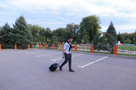 In Parking Goes Young Man Arabic Attractive Appearance With Beard and Dark Hair Dressed in Suit Black Pants White Shirt With Black Sweater on Shoulders and Roll Black Suitcase Laggage in His Hand. Concept of Business Trip, Business Meeting, Travel, Busineの写真素材