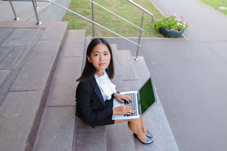 Modern young women, business people, international students holds laptop and study work plan, solve important problems and smiling sits on stairs on background of gray wall of business center outdoors. Woman with black hair, dressed in white blouse and clの写真素材